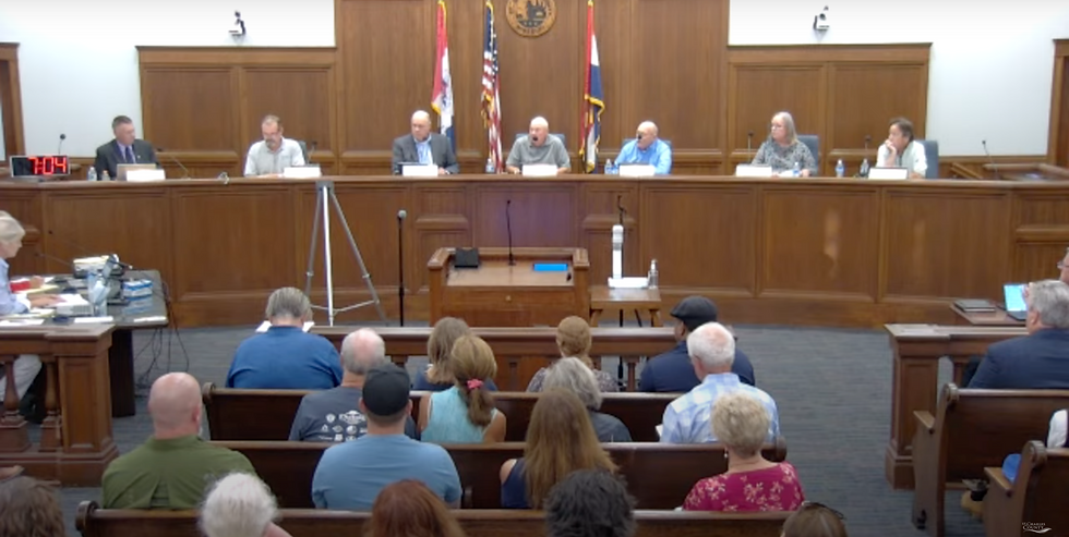 A panel sits at a wood-paneled desk in a courtroom, addressing an audience. A clock reads 7:04. Flags and microphones are present.