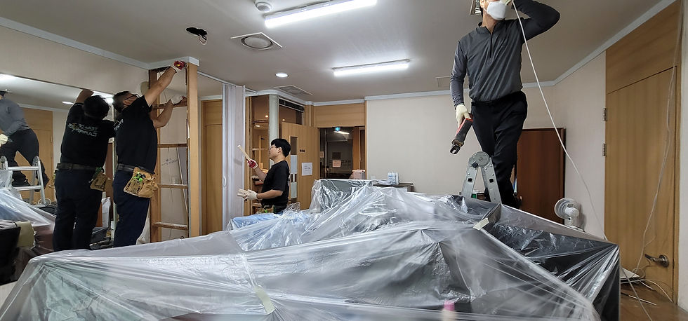 비닐로 덮은 방 안에서 작업복을 입은 남성 4명이 사다리와 전동공구로 벽과 천장을 수리하는 공사 현장