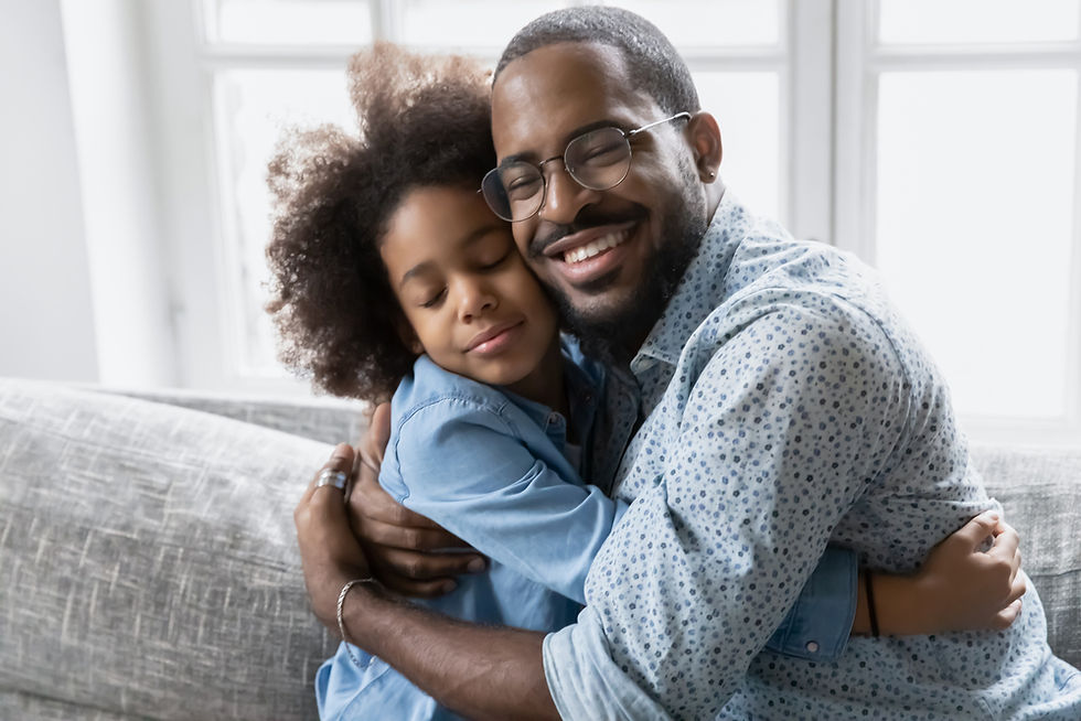 Father and daughter hugging warmly on a gray couch, both wearing blue shirts. A bright window is in the background, creating a cozy mood.