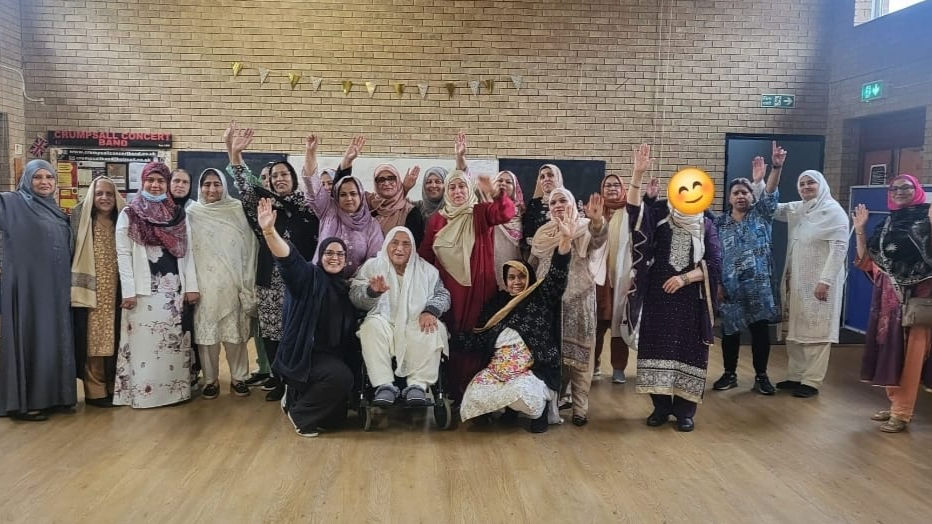 South Asian women of all ages, in traditional dress, stand together in a large hall. They smile and wave at the camera.