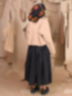 A person with intricate braided hair stands in a room of hanging paper patterns, wearing a tan sweater, black skirt, and black shoes.