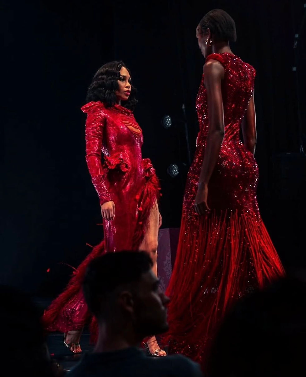 Two models in elegant red dresses walk a dimly lit runway, creating a dramatic and glamorous atmosphere.