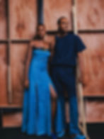 Two models in blue outfits stand against a wooden backdrop. One wears a dress with a slit, the other in loose pants. They look confident.