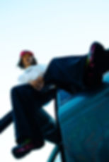 Person in a white shirt and red cap leans out of a car door, relaxed posture. Bright sneakers stand out, sky visible in the background.