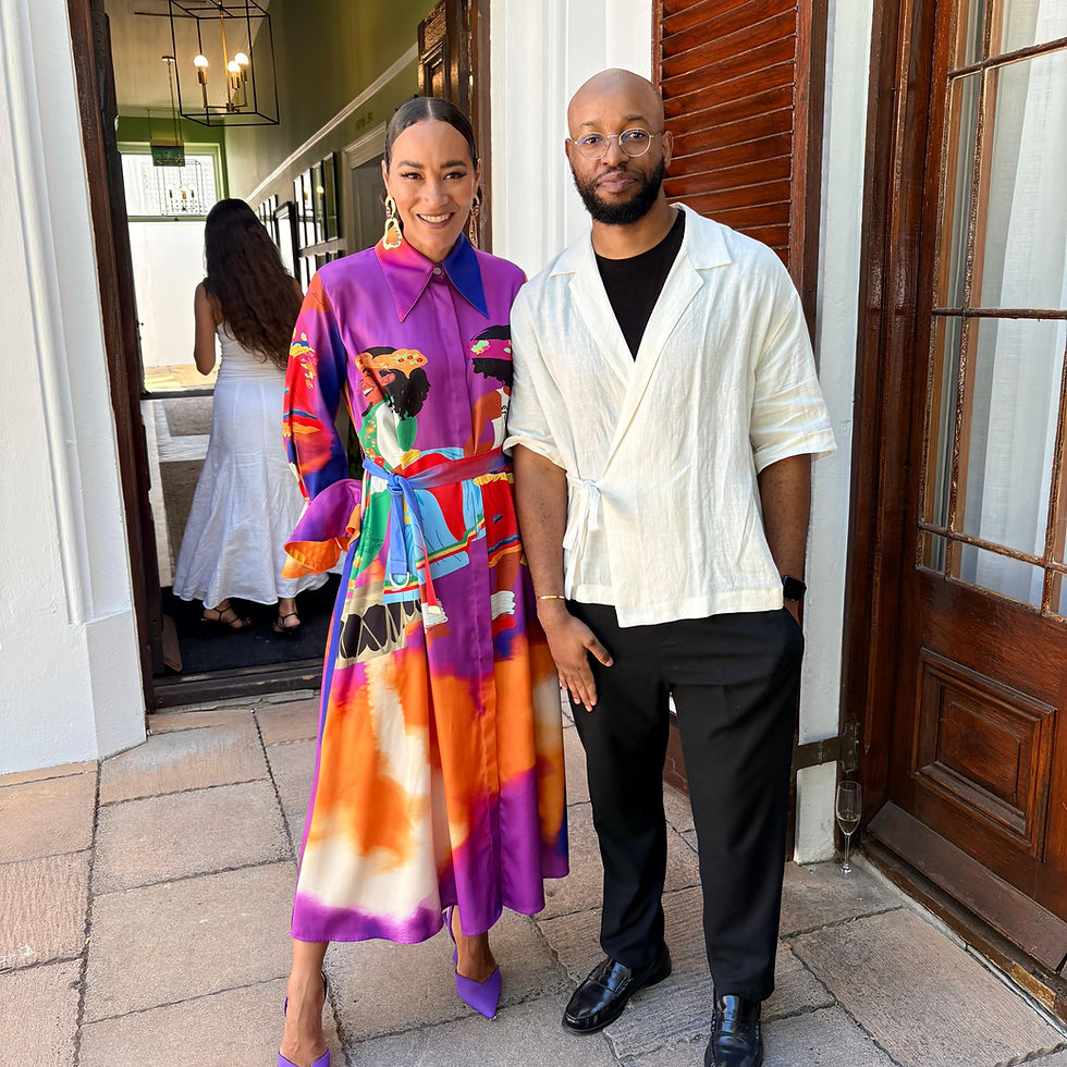 A woman in a vibrant purple and orange dress and a man in a white shirt pose smiling outside a building with wooden doors.