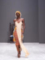 Model in a striped dress with fringe details and a headband walks on a white runway, gray background. Mood is confident and stylish.