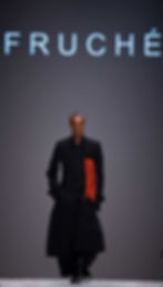 Model walks runway in black coat with red bead details. Background text reads "FRUCHÉ." Gray backdrop, confident mood.
