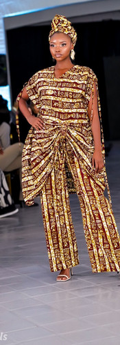 Model in kante jumpsuit with black accents walks runway wearing red visor. Brightly lit venue, audience in background, stylish atmosphere.