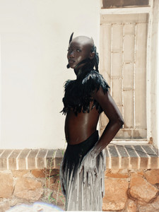 Person in feathered top and flowing skirt poses confidently outside a rustic hut with straw roof and brick foundation, in a sunlit garden.