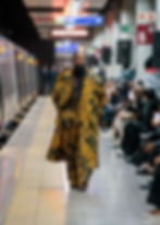 Man in a patterned yellow outfit walks on a subway platform runway, surrounded by seated crowd. Train on left, exit signs above. Fashion show vibe.