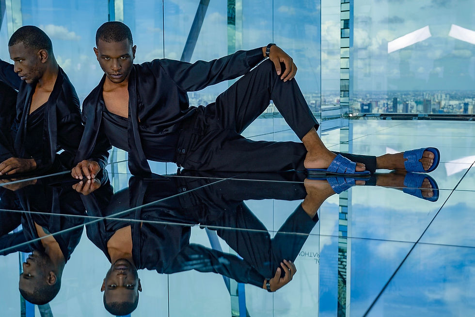 Two men in black outfits pose on a reflective glass floor, one reclining in blue sandals, with a city skyline behind them.