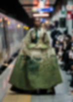 Model in green patterned dress walks confidently on a subway platform runway. Stylish crowd views under bright station lights.