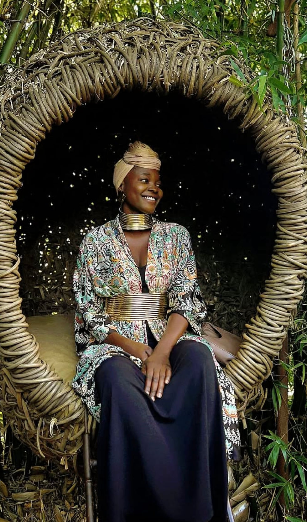 Smiling woman in a colorful outfit sits on a woven swing in a bamboo setting, exuding a joyful mood. The swing is circular and rustic.