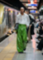Model in white blouse and green pants walks on subway platform, holding a patterned bag. Train in background, onlookers seated nearby.