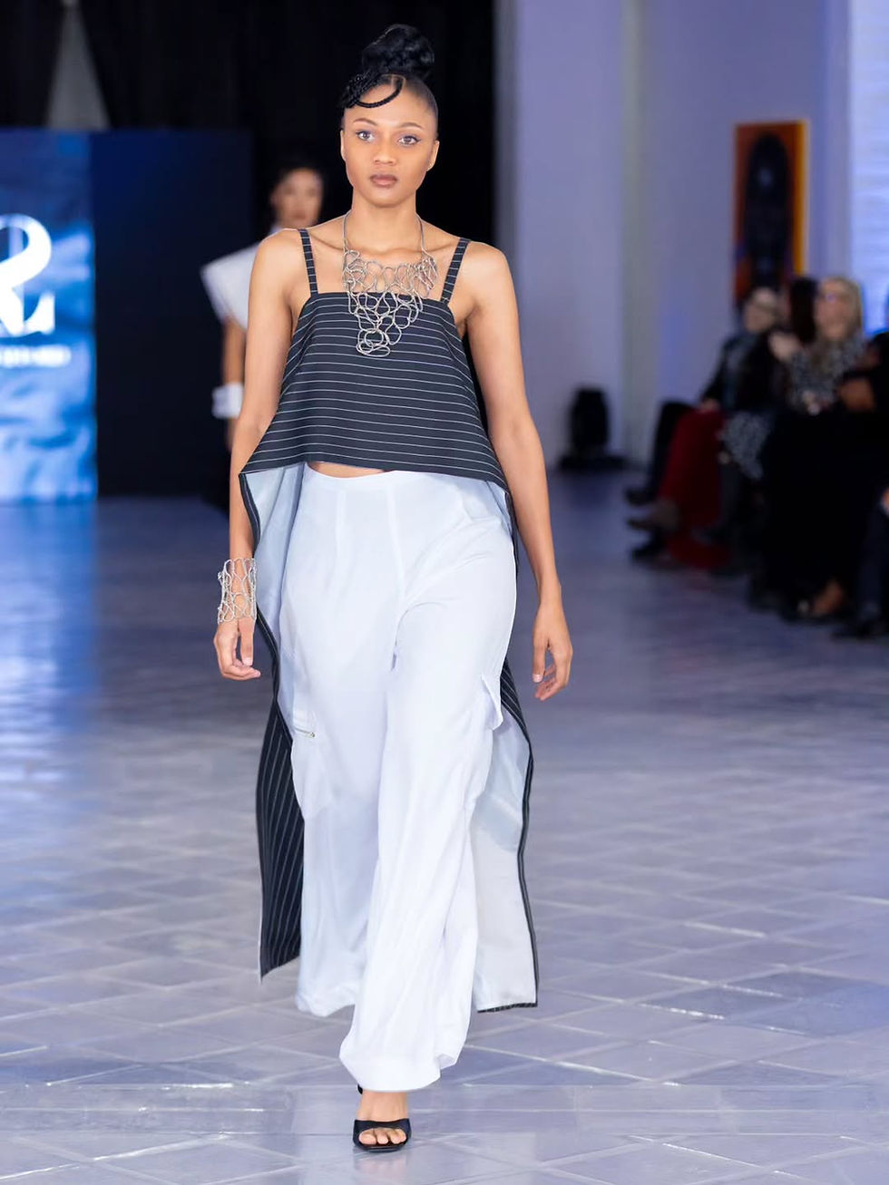 Model on a runway in a striped top, white pants, black heels, and silver jewelry. Bright background with audience seated on the side.