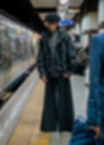 Person in a black jacket and wide pants stands on a train platform. Background shows a train and seated passengers. Modern, urban setting.