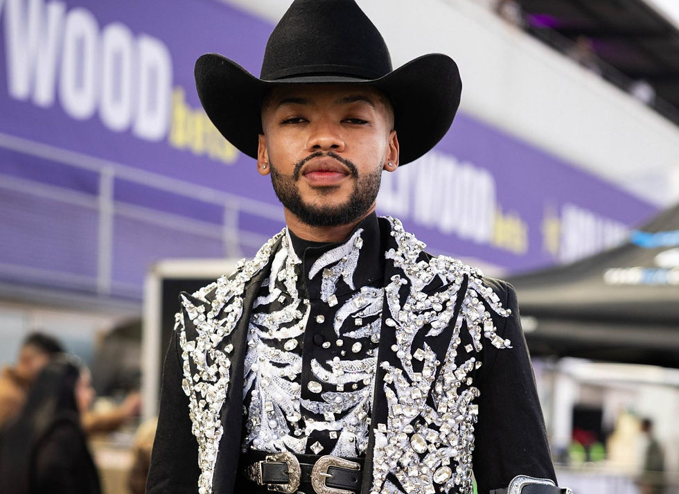 Man in a black cowboy hat and jeweled black suit stands at an event, with a purple backdrop and blurred crowd.