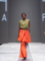 Model walks runway in orange skirt and green mesh top, against gray backdrop. Confident expression, minimalist fashion show setting.