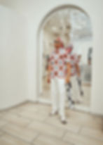 Man in a red and white patterned outfit stands confidently in front of a mirror with white quilted pants, sunglasses, and sandals.