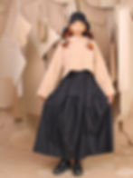 A woman poses in a beige sweater and black skirt with braids adorned with orange accents. Background features hanging paper sheets.