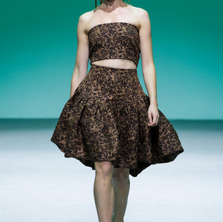 Model in patterned dress and top outfit and white sleaves walks runway. Infront of Green backdrop.