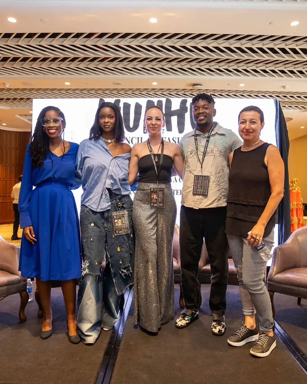 Five people pose smiling on stage with a geometric ceiling above. They wear colorful outfits, and a large screen displays blurred text.