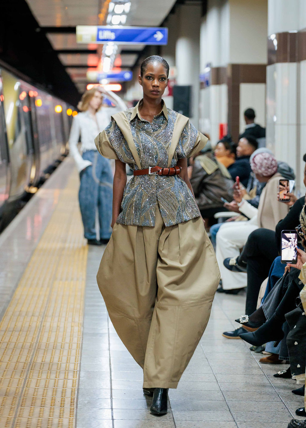 Model in cream pants and cream and grey patterned shirt walks confidently on a subway platform runway. Stylish crowd views under bright station lights.