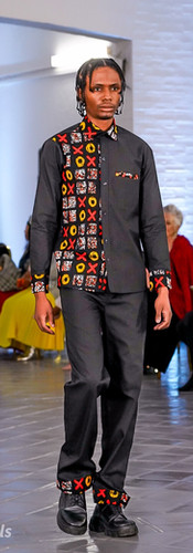 Model in grey and floral suit with black accents walks runway wearing red visor. Brightly lit venue, audience in background, stylish atmosphere.