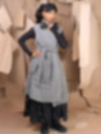 Woman with braided hair in a gray wool dress poses confidently against a backdrop of hanging craft paper. Wears black shoes and floral earrings.