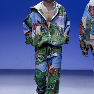Model in green and blue, fringe-detailed outfit walks on a fashion runway against a teal background, exuding confidence and style.
