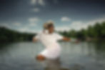 A person in a white dress poses dynamically in a lake, wearing large earrings and multiple bracelets, with a forest and cloudy sky behind.