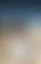 Person with red-painted skin and ornate headpiece poses in rocky, grassy landscape under blue sky, wearing a white dress and bead necklaces.