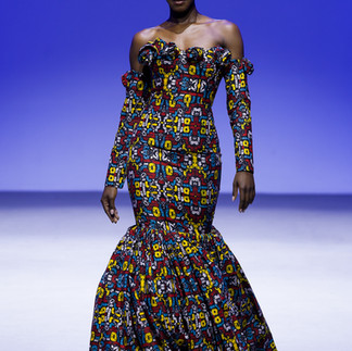 Model in pattened green yellow and red dress outfit and white sleaves walks runway. Infront of blue backdrop.