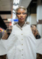 A person stands in a subway, wearing a white outfit with text patterns. Gold hair accessory and bold eyelashes enhance their confident look.