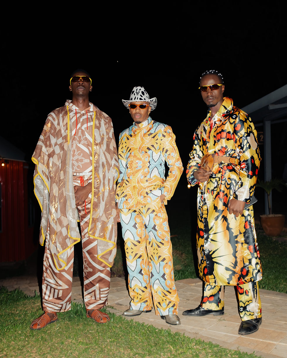 Three men pose at night outdoors in bold patterned outfits, sunglasses and a white cowboy hat, on a patio with grass and dark sky.