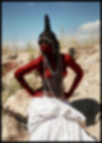 Person with red-painted skin, adorned in beads and shells, poses confidently against a rocky, grassy landscape under a blue sky.