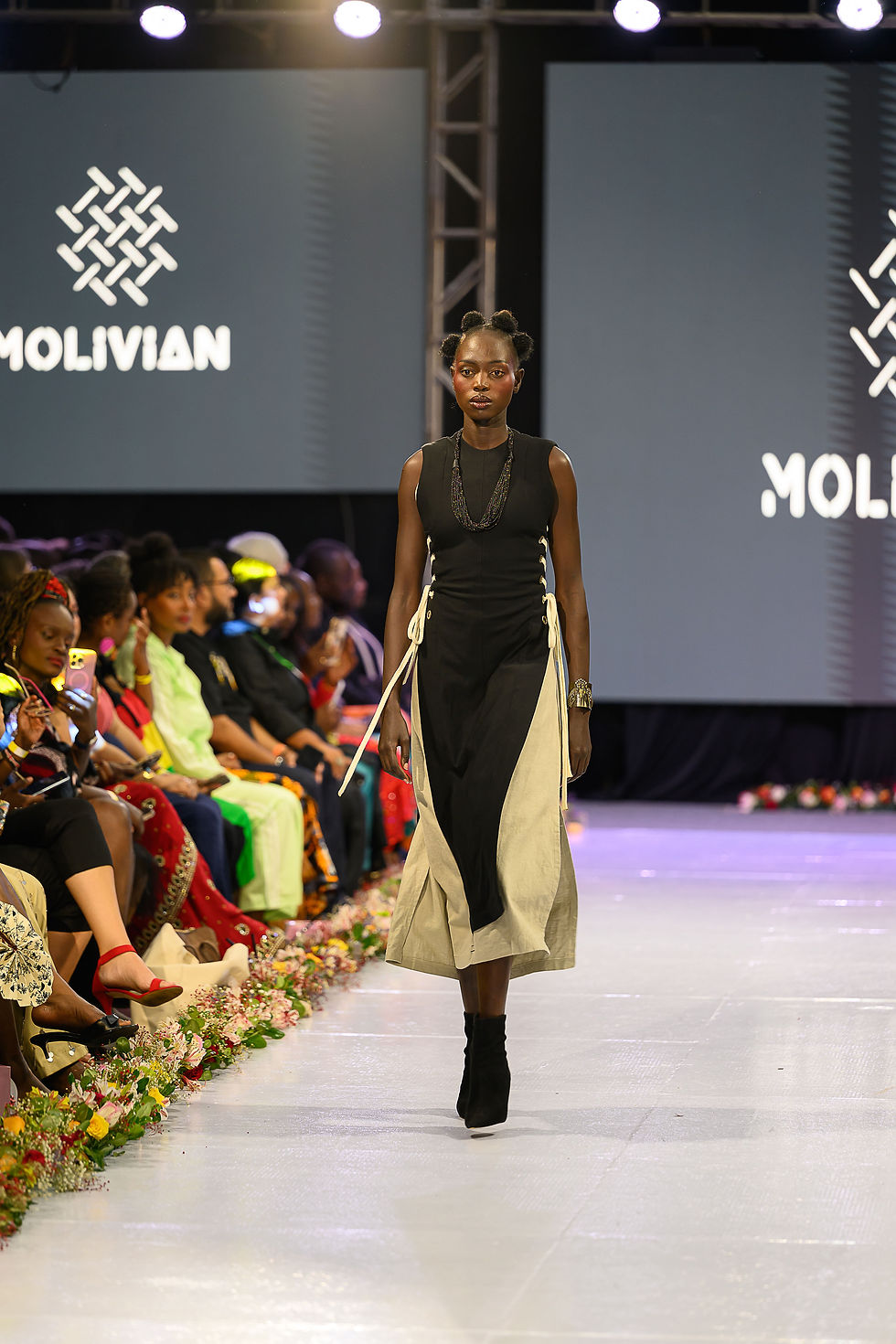 Model walking down a runway in a black and beige dress, surrounded by seated audience. Stage lights and "MOLIVIAN" text visible.