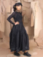 A woman in a sheer black top and a long black skirt with white specks poses confidently. She stands against a background of hanging paper patterns.