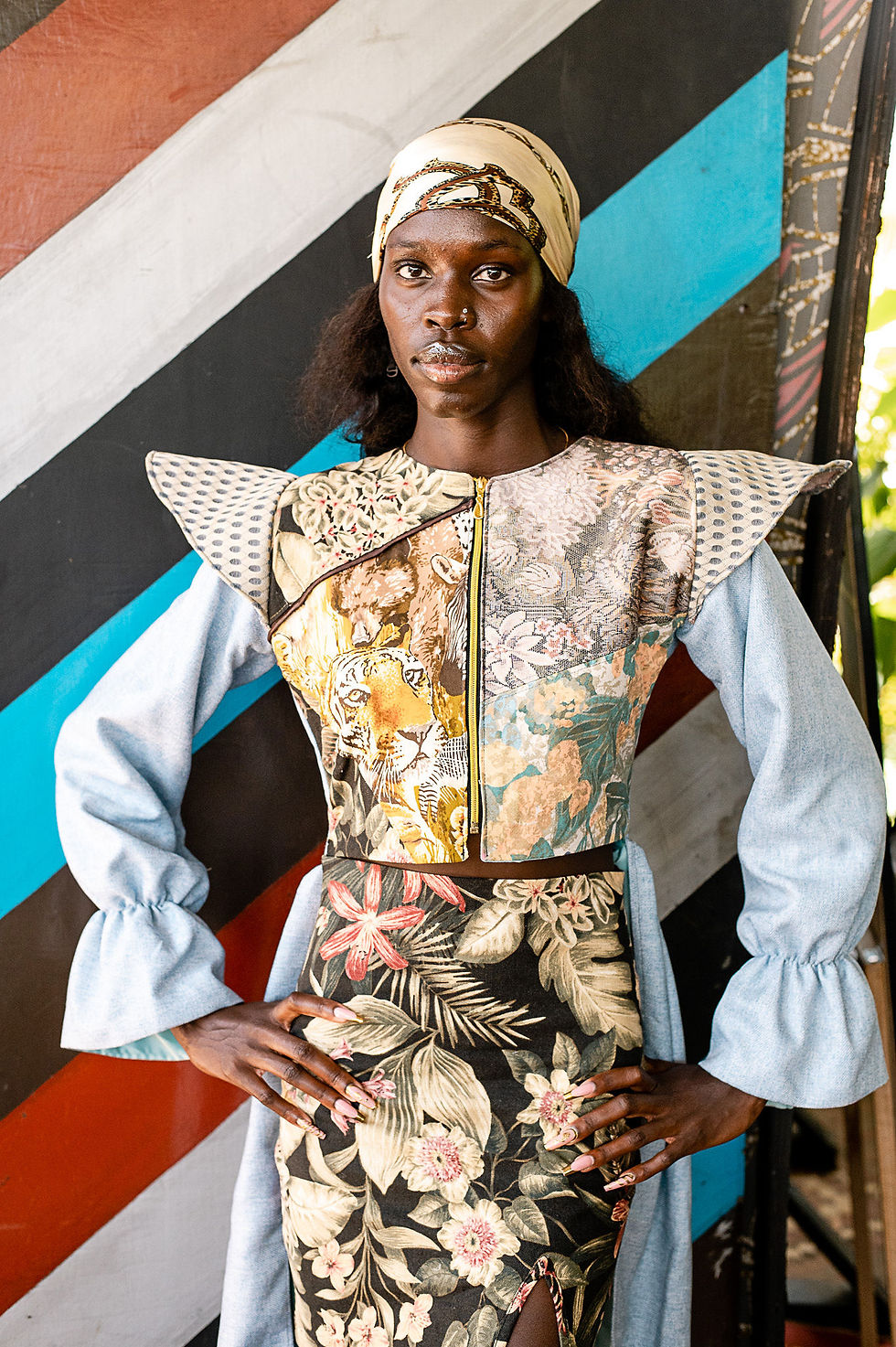 A person poses confidently in patterned clothing against a colorful striped background, wearing a headscarf. The mood is bold and stylish.