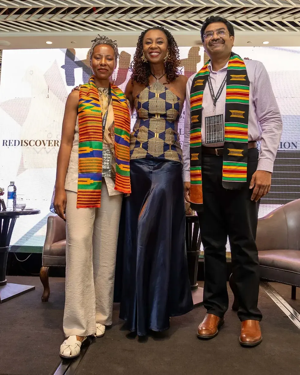 Three people wearing colorful kente stoles stand smiling on stage. The background features a patterned wall and the word "REDISCOVER."