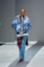 Model walking on runway in blue camo jacket, plaid skirt, ripped jeans, and red shoes. Gray background, confident expression.