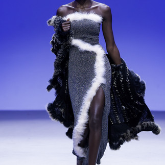 Model in gray and white outfit walks runway. Infront of blue backdrop.