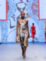 Model walks runway in colorful patterned dress and red headband, with vibrant abstract backdrop. Confident and stylish mood.