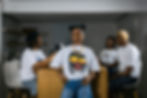 Four people in a modern kitchen. One poses confidently in a graphic tee, while three converse casually in the background. Minimalist decor.