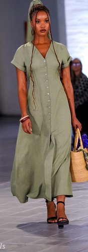 Model in green dress with black accents walks runway wearing red visor. Brightly lit venue, audience in background, stylish atmosphere.