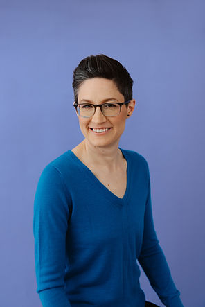 Danika, a white person with short brown hair, smiles at the camera. They are sitting and wearing a turquoise sweater.