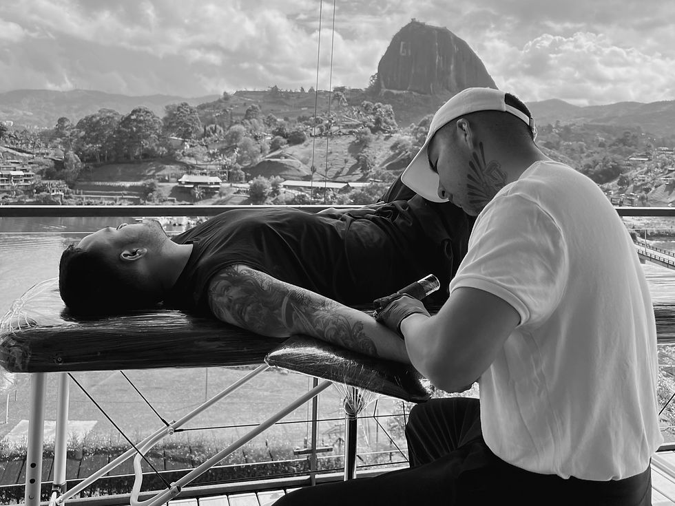 A skilled tattoo artist named Apz is shown in the process of tattooing. The backdrop of the image features the majestic "El Peñón de Guatapé" located in the picturesque region of Antioquia, Colombia.