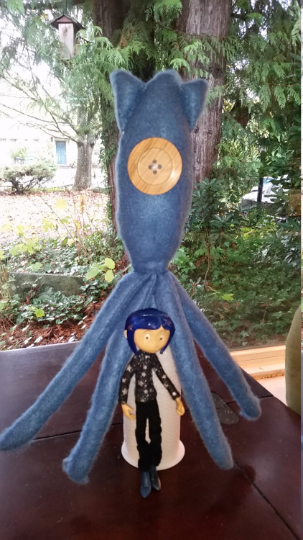 a blue squid-looking toy made of felt with a large button where the eyes should be, stands over a toy figure of Coraline