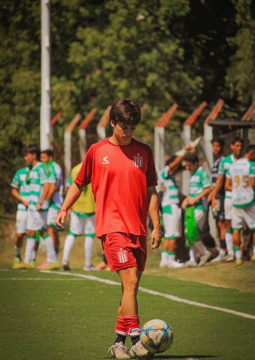 A soccer player in a red kit dribbles a ball on a grassy field. Opponents in green and white jerseys are in the background, next to a fence.