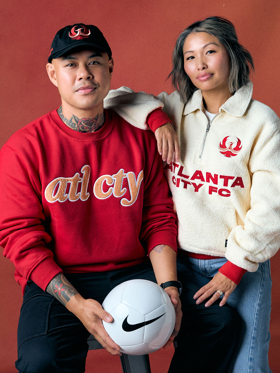 Two Atlanta City FC supporters pose against a red backdrop, holding a white Nike soccer ball, wearing red and cream team gear.